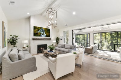 Open-concept living room with vaulted ceiling, white brick fireplace, and floor-to-ceiling windows overlooking wooded area