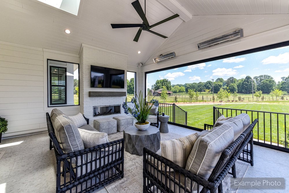 Covered patio with wicker seating, brick fireplace, ceiling fan, wall-mounted TV, and open views of a green backyard.