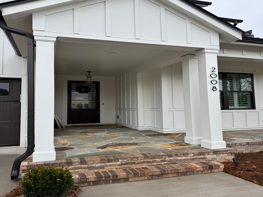 White farmhouse front porch with columns, black door, stone flooring, and brick steps