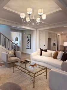 Modern living room with tray ceiling, white chandelier, and neutral furniture with staircase visible