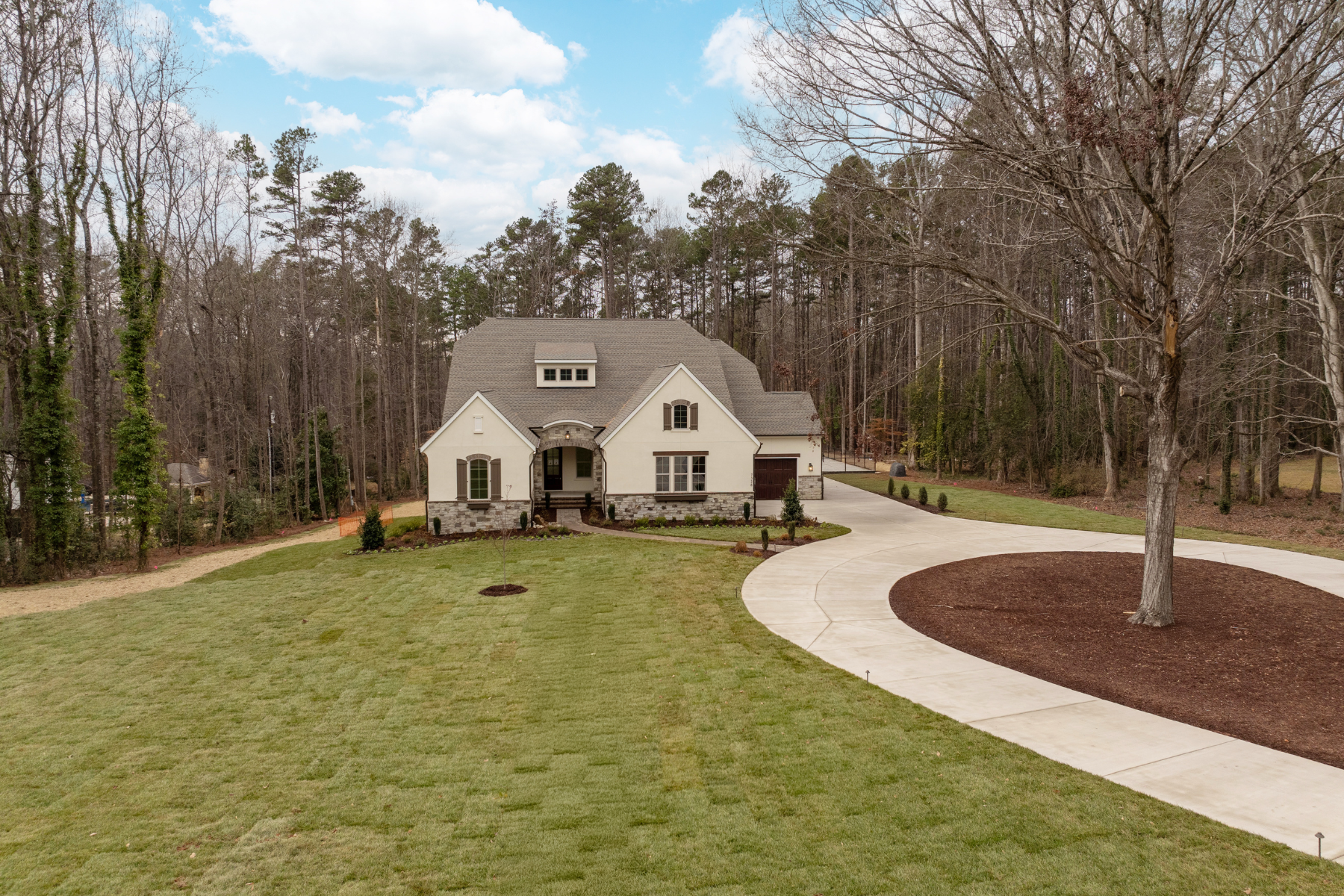 Single-story home with cream stucco exterior, stone accents, and curved driveway on wooded lot