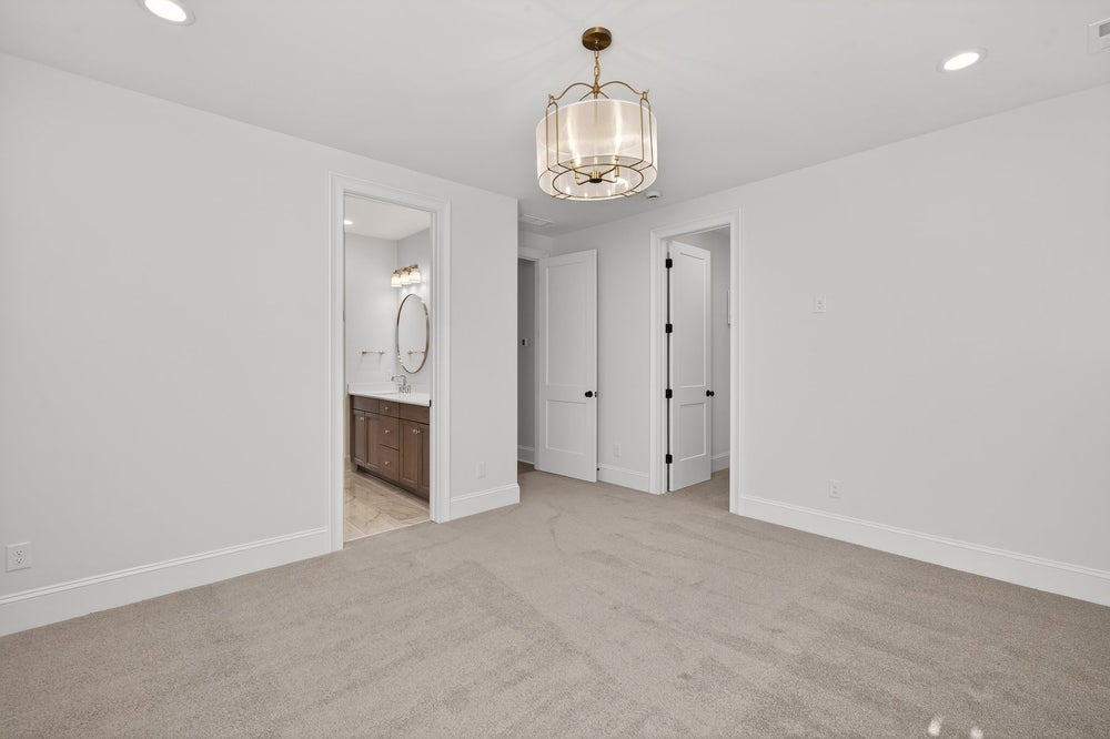 Empty bedroom with beige carpet, decorative light fixture, and ensuite bathroom