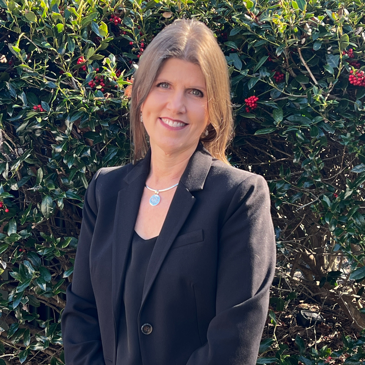 Professional headshot of woman in black blazer outdoors with greenery background