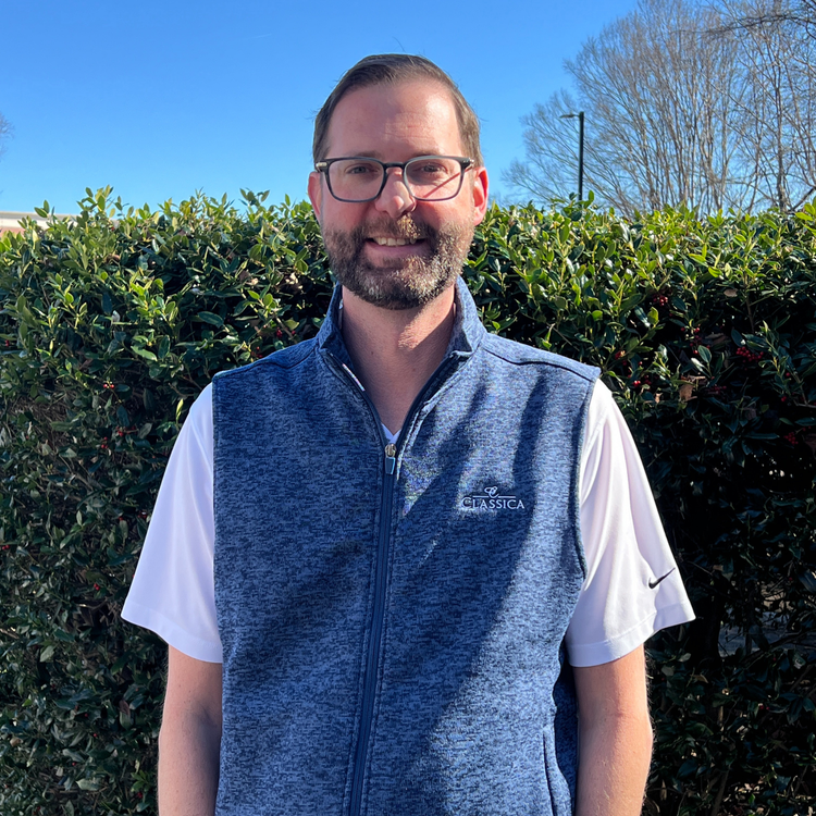 Man wearing glasses and blue Classica vest standing outdoors with hedge background