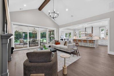 Open-concept living room with vaulted ceiling, wood beams, and kitchen visible in background, with sliding doors to patio