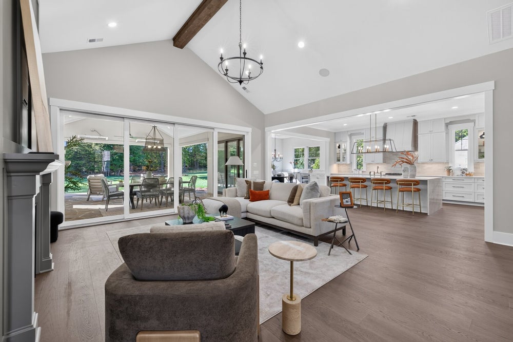 Open-concept living room with vaulted ceiling, wood beams, and kitchen visible in background, with sliding doors to patio