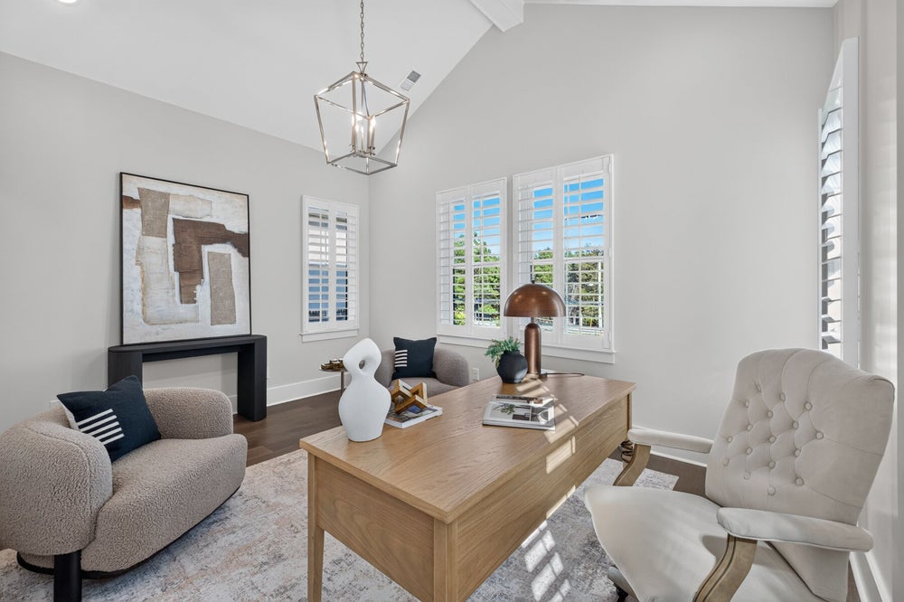 Home office with vaulted ceiling, wood desk, tufted chair, and plantation shutters on multiple windows