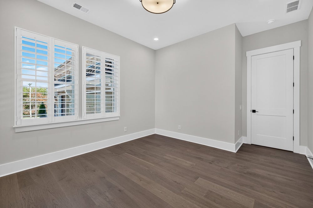 Empty bedroom with dark hardwood floors, gray walls, and white plantation shutters on corner windows.