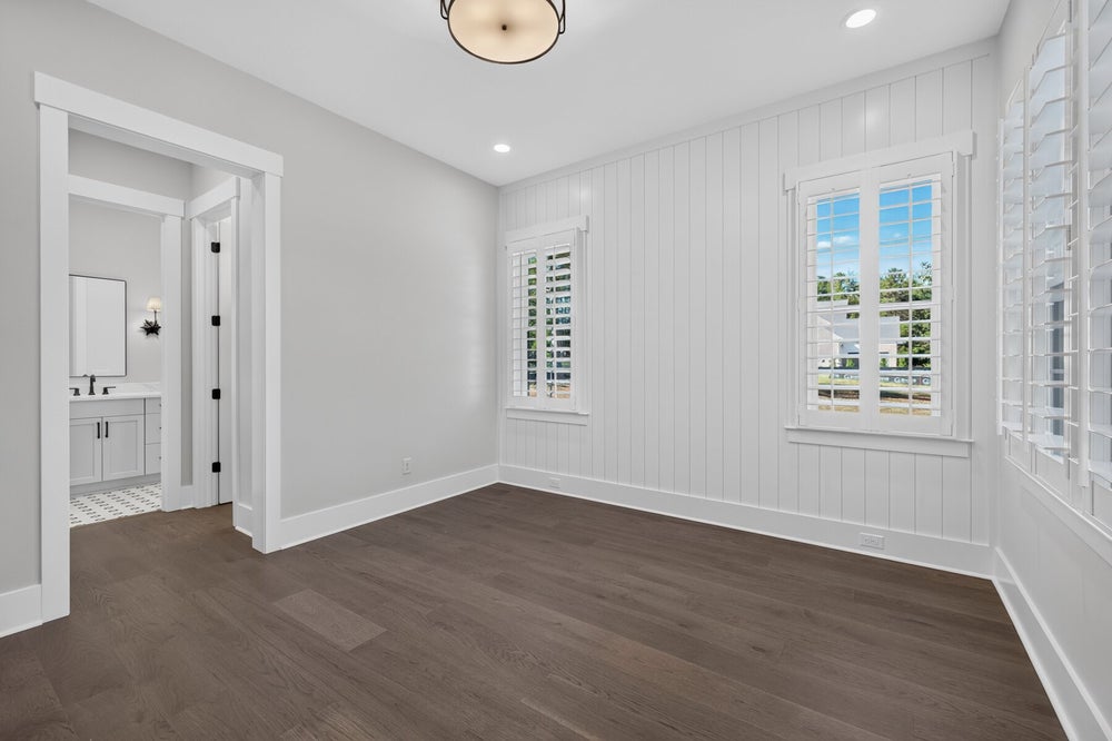 Empty bedroom with dark hardwood floors, shiplap accent wall, plantation shutters, and ensuite bathroom access.