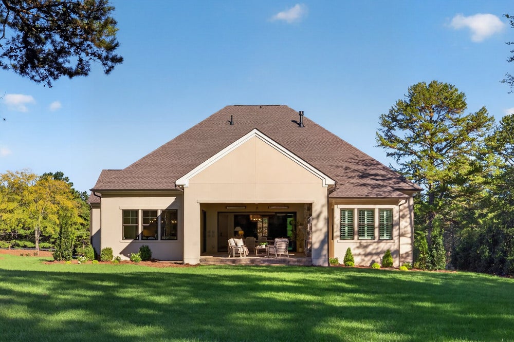 Rear view of single-story home with covered patio, seating area, and large green backyard surrounded by trees.
