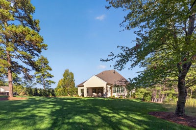 Single-story home with tan exterior and covered patio, set on a large grassy lot surrounded by mature trees.