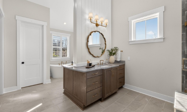 Spacious master bathroom with double vanity, round mirror, gold light fixture, and freestanding soaking tub in background.
