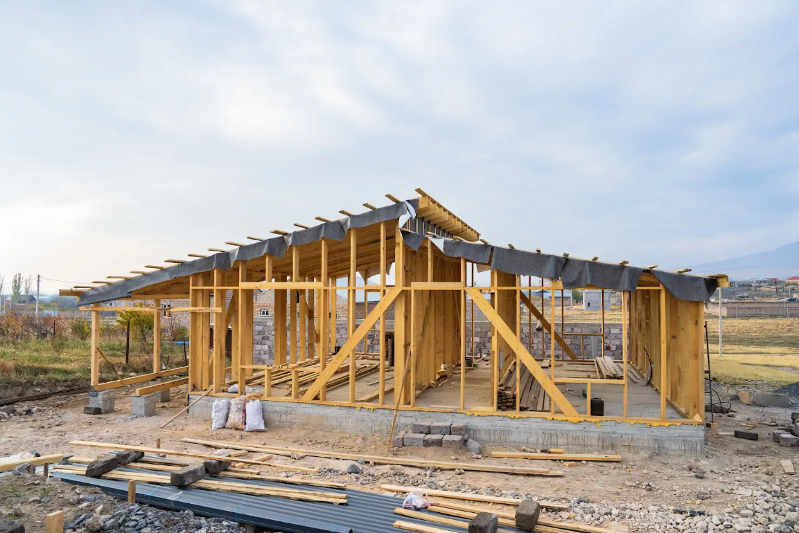 Wood-framed house under construction with exposed studs and roof trusses on concrete foundation