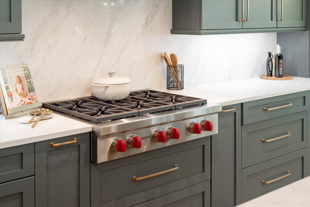 Kitchen with gray-green cabinets, white quartz countertops, marble backsplash, and professional gas range with red knobs