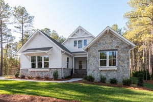 Craftsman-style home with white shingle siding, stone facade, covered front porch, and landscaped yard surrounded by pine trees.