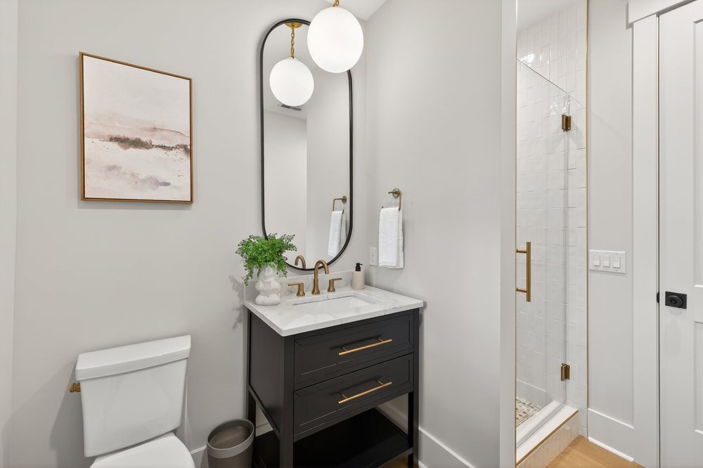 Modern bathroom with dark vanity, marble countertop, oval mirror, and glass shower enclosure with brass fixtures