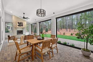 Covered patio with dining table, stone fireplace, and large windows overlooking wooded backyard
