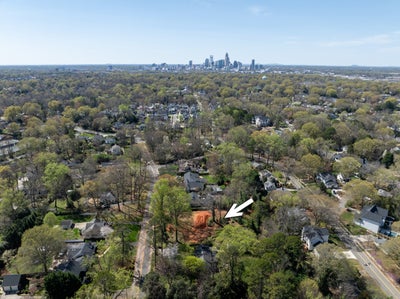 Aerial view of a tree-lined residential neighborhood with a city skyline visible in the background and an arrow marking a lot.