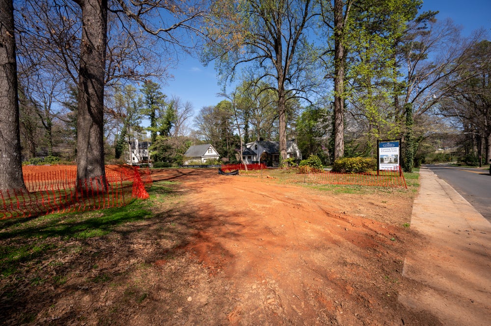 Vacant residential lot with orange safety fencing and construction sign in tree-lined neighborhood