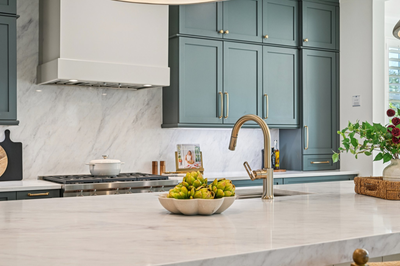 Kitchen with teal-green cabinets, marble countertops and backsplash, gold faucet, and stainless steel range with white hood