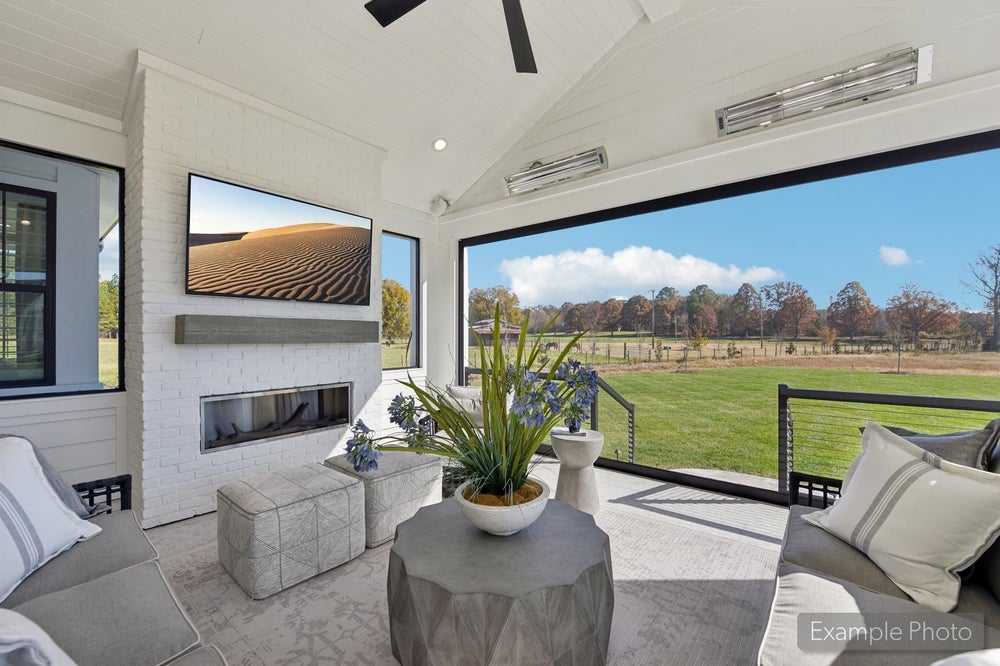 Covered outdoor patio with brick fireplace, wall-mounted TV, ceiling fan, and open view of green pasture