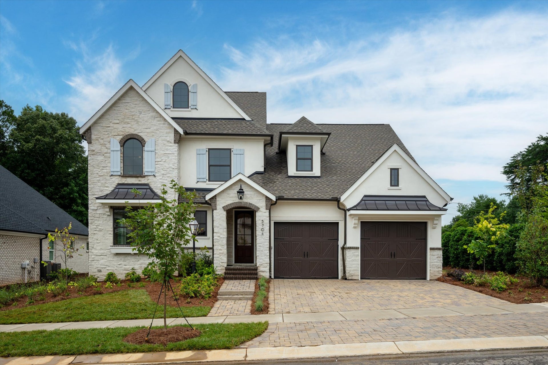 Two-story home with white stone exterior, dark roof, two-car garage, and paver driveway