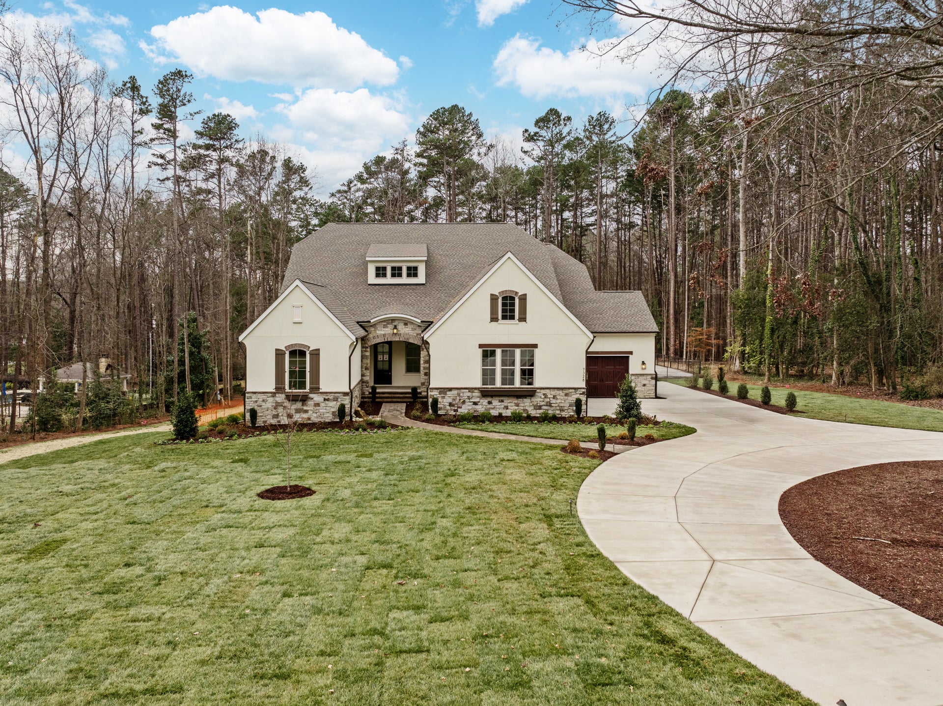 White stucco home with stone accents, arched entry, single-car garage, curved driveway, and wooded lot