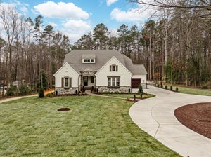 White stucco home with stone accents, arched entry, single-car garage, curved driveway, and wooded lot