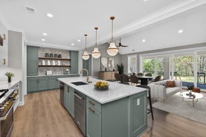 Open-concept kitchen with sage green cabinets, marble island, and pendant lights, flowing into dining and living areas with large windows.