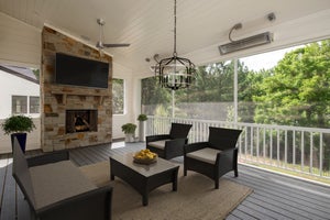 Covered porch with stone fireplace, mounted TV, dark wicker seating, and screened views of trees