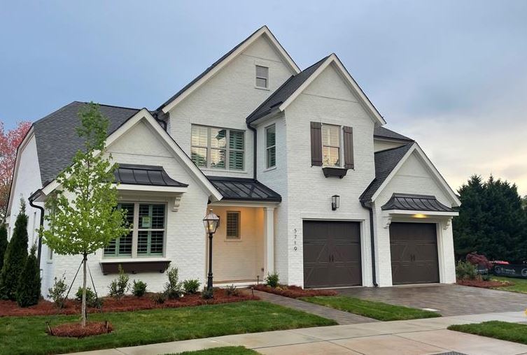 Two-story white brick home with dark shutters, metal roof accents, and two-car garage