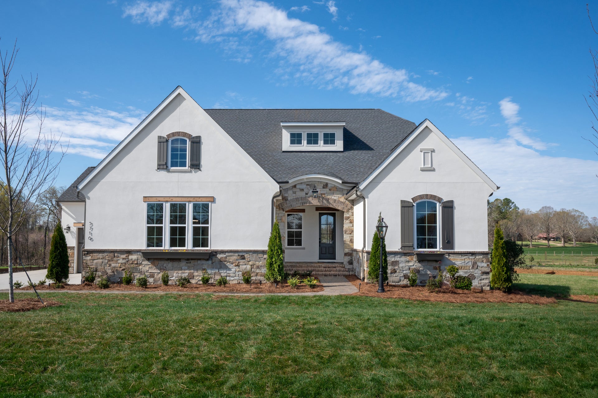 Single-story home with white stucco and stone exterior, arched entry, gray shutters, and manicured front lawn