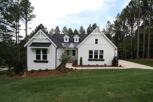Modern farmhouse with white board-and-batten siding, dark roof, and concrete driveway surrounded by trees