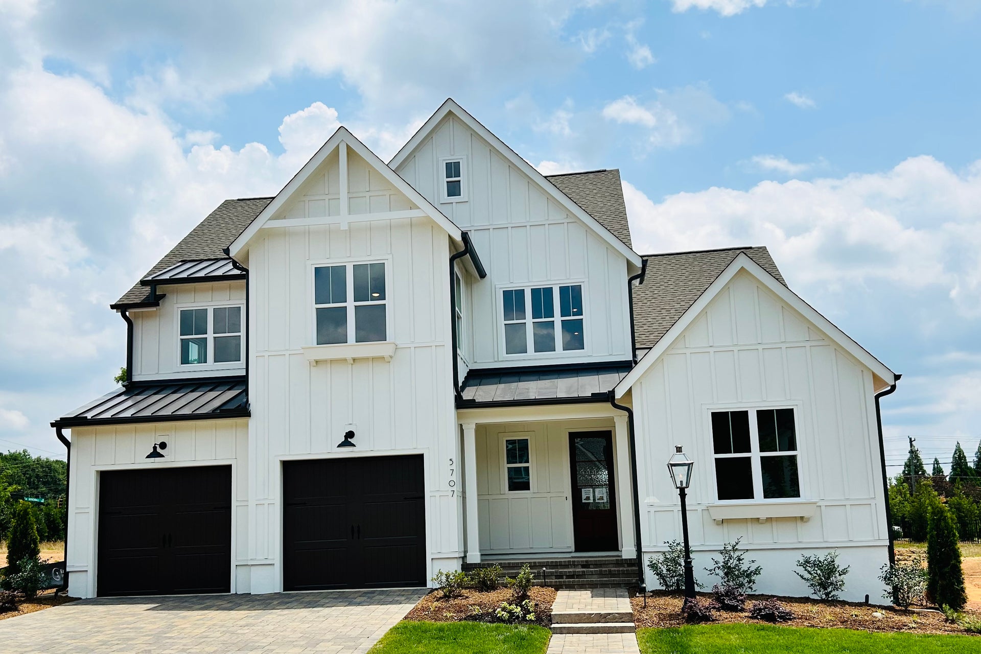 Two-story white farmhouse with board-and-batten siding, black metal roof accents, and two-car garage