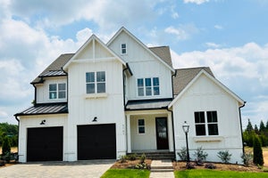 Two-story white farmhouse with board-and-batten siding, black metal roof accents, and two-car garage