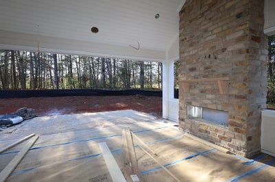 Covered outdoor patio under construction with tall stone fireplace, protected floors, and wooded backyard view