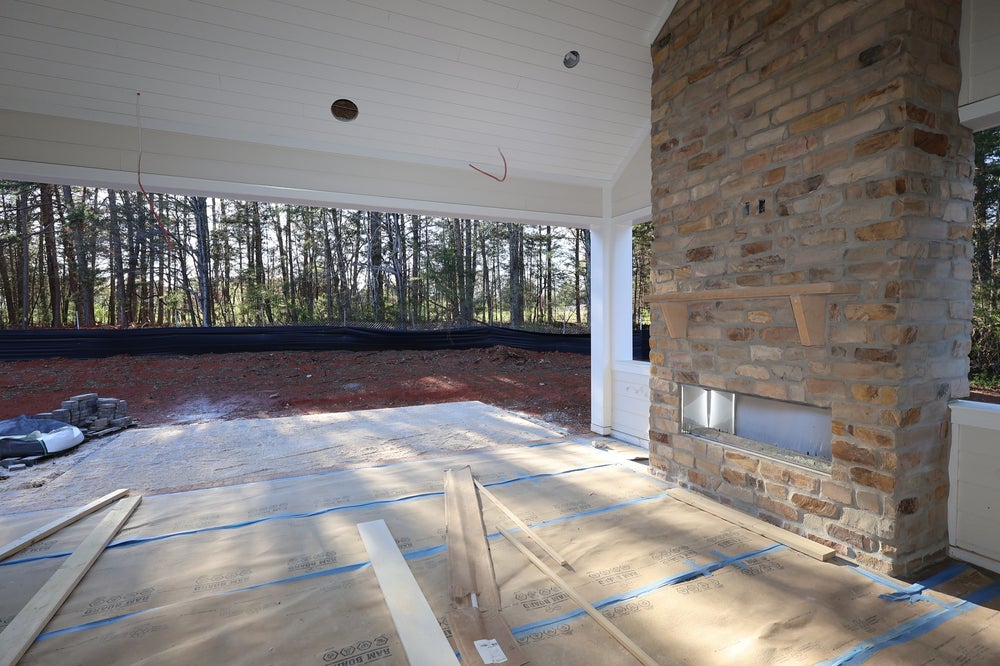 Covered outdoor patio under construction with tall stone fireplace, protected floors, and wooded backyard view