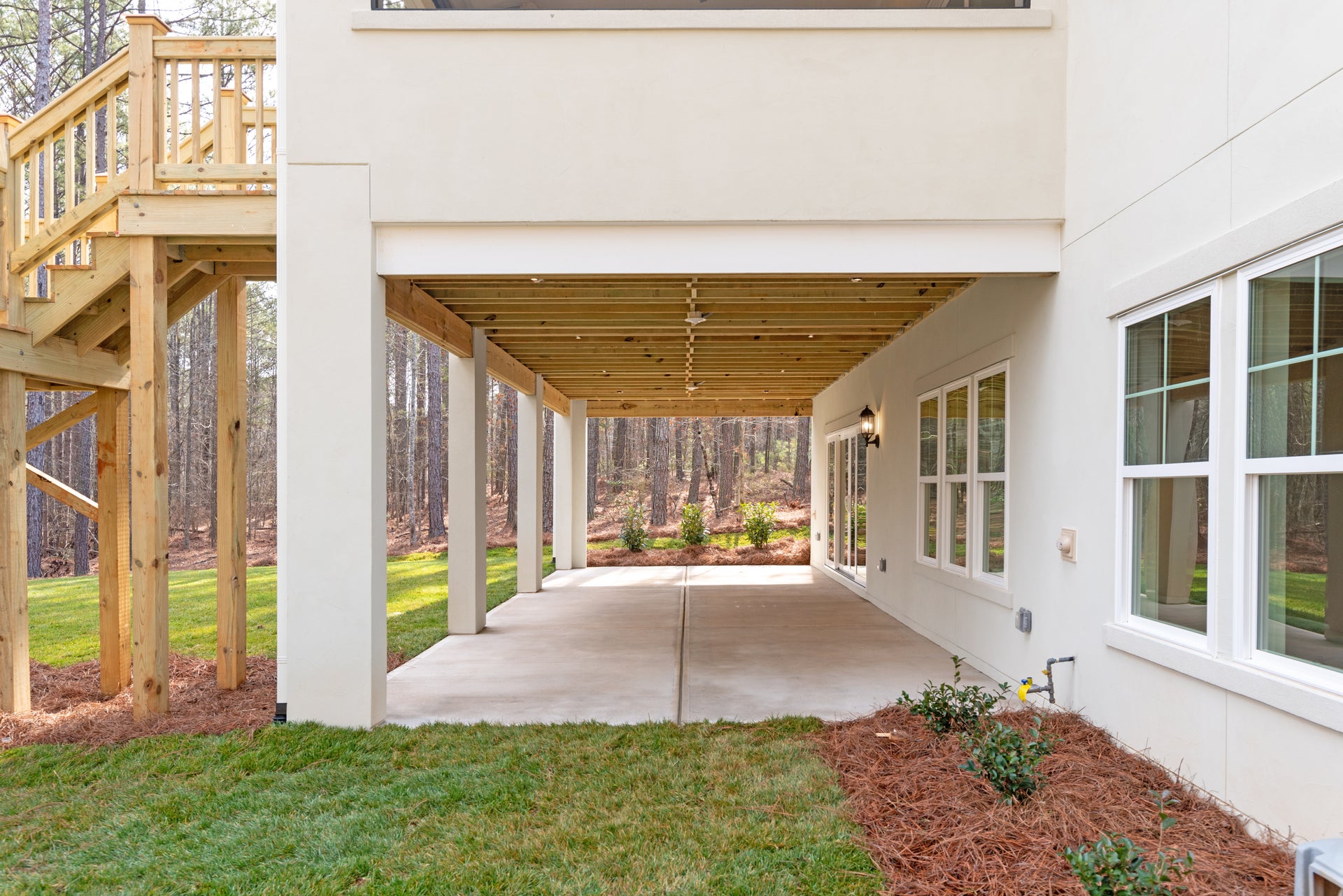 Covered concrete patio under elevated deck with wood ceiling, ceiling fan, and wooded backyard view