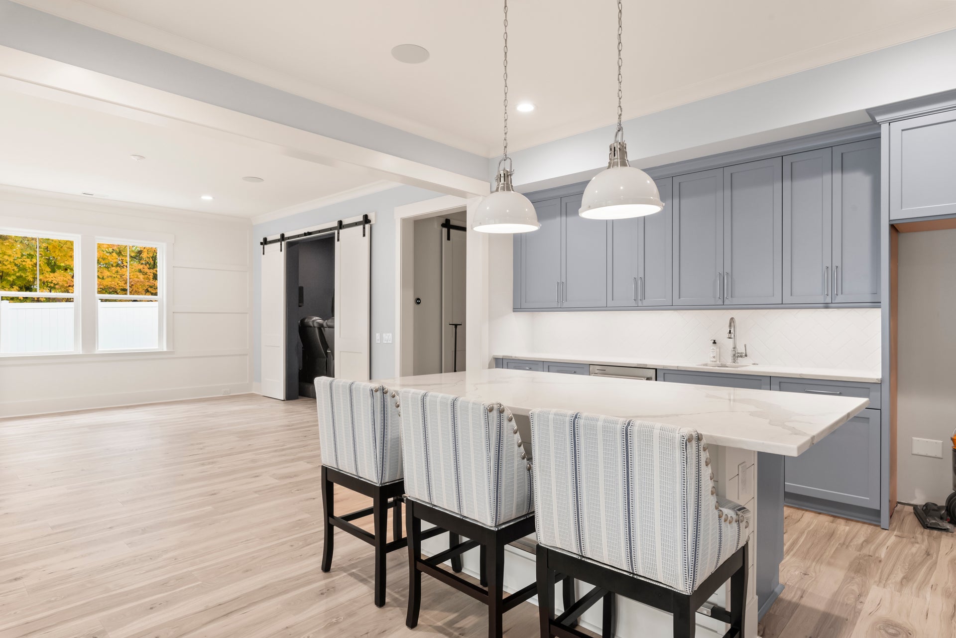 Kitchen with gray cabinets, white island, three bar stools, pendant lights, and barn doors on hardwood floors
