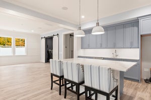 Kitchen with gray cabinets, white island, three bar stools, pendant lights, and barn doors on hardwood floors