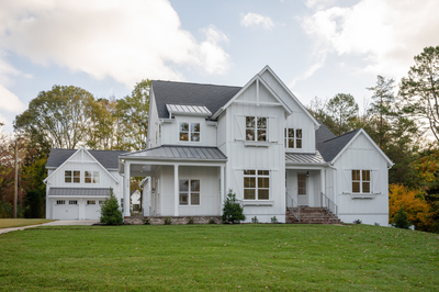 Two-story white farmhouse with board and batten siding, metal roof accents, covered porch, and attached two-car garage on a green lawn.