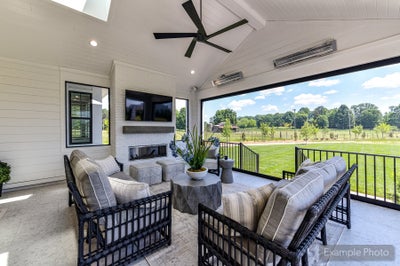 Covered patio with wicker seating, brick fireplace, ceiling fan, wall-mounted TV, and open views of a green backyard.