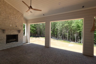 Covered outdoor patio with stone fireplace, ceiling fan, brick floor, and open views of wooded backyard