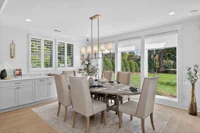 Dining room with oval wood table, six upholstered chairs, brass chandelier, and large windows overlooking a backyard
