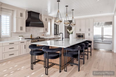 Large kitchen island with white countertop, black barstools, pendant lights, and cream cabinets with black range hood
