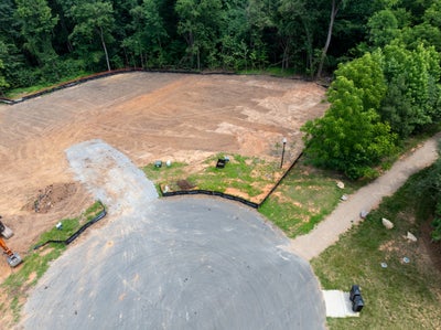 Aerial view of a cleared and graded residential building lot with silt fencing, gravel driveway, and wooded border.