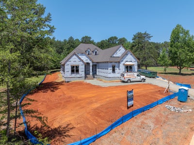 New home under construction with house wrap exterior, gray roof, and graded red clay lot surrounded by trees