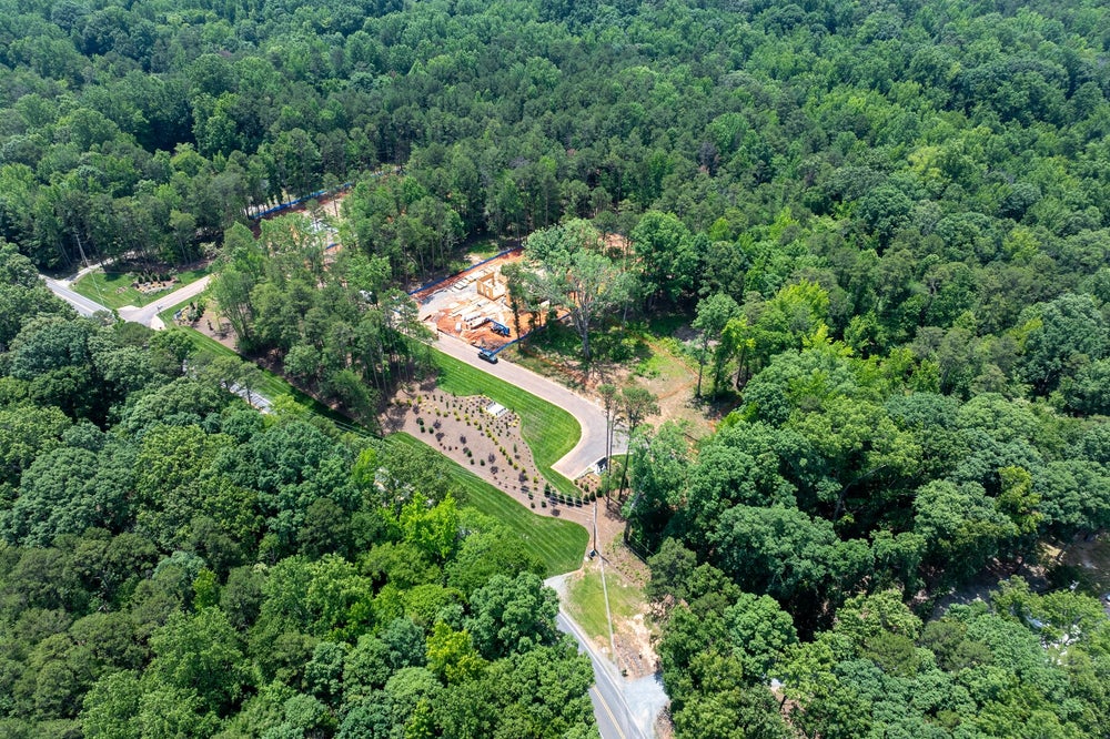 Aerial view of a wooded residential development with a home under construction and a winding road through dense trees.