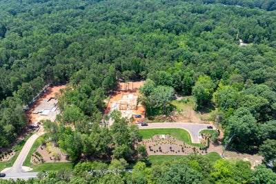 Aerial view of new home construction site with concrete foundations surrounded by dense trees and landscaped entrance road.