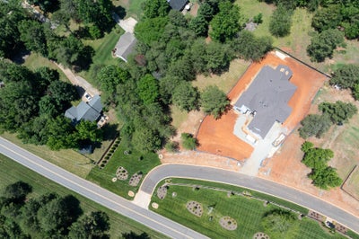 Aerial view of a home under construction with exposed red clay lot, curved driveway, and surrounding trees